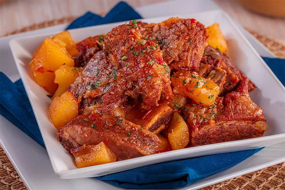 costela com mandioca dentro de um prato fundo branco de porcelana em cima de um pano azul escuro e dentro de outro prato branco de porcelana