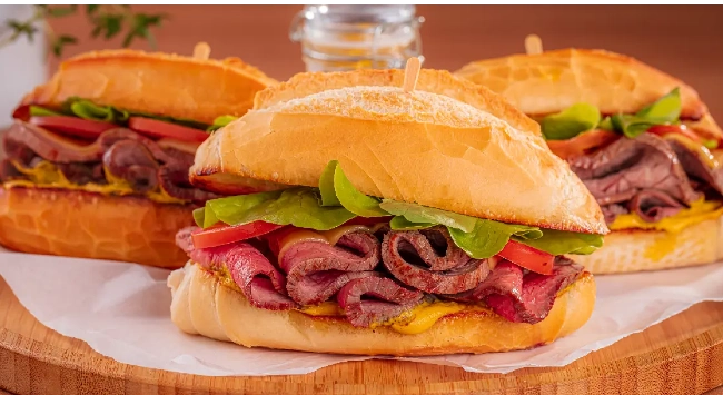 Sanduíches de rosbife suculentos em pães tipo baguete, posicionados sobre uma tábua de madeira. 
